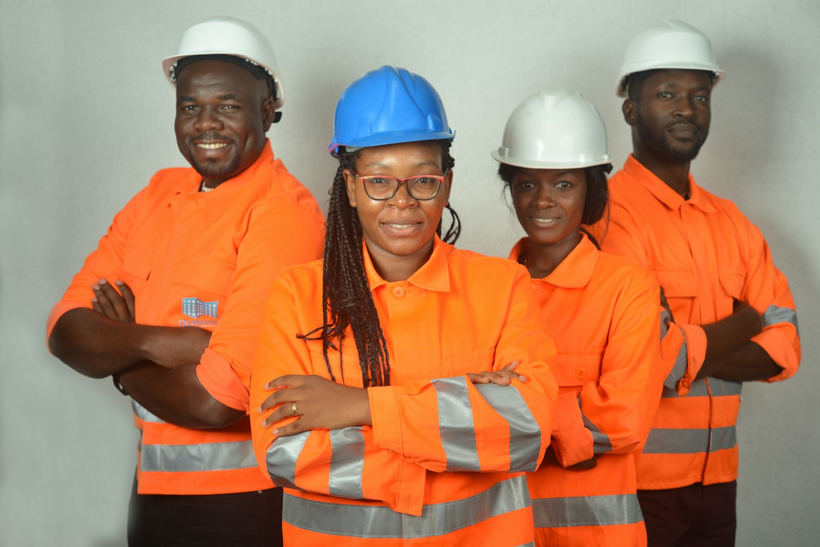 Four workers in orange hi-vis jackets with reflective bands and hard hats posing with arms crossed against a plain studio backdrop