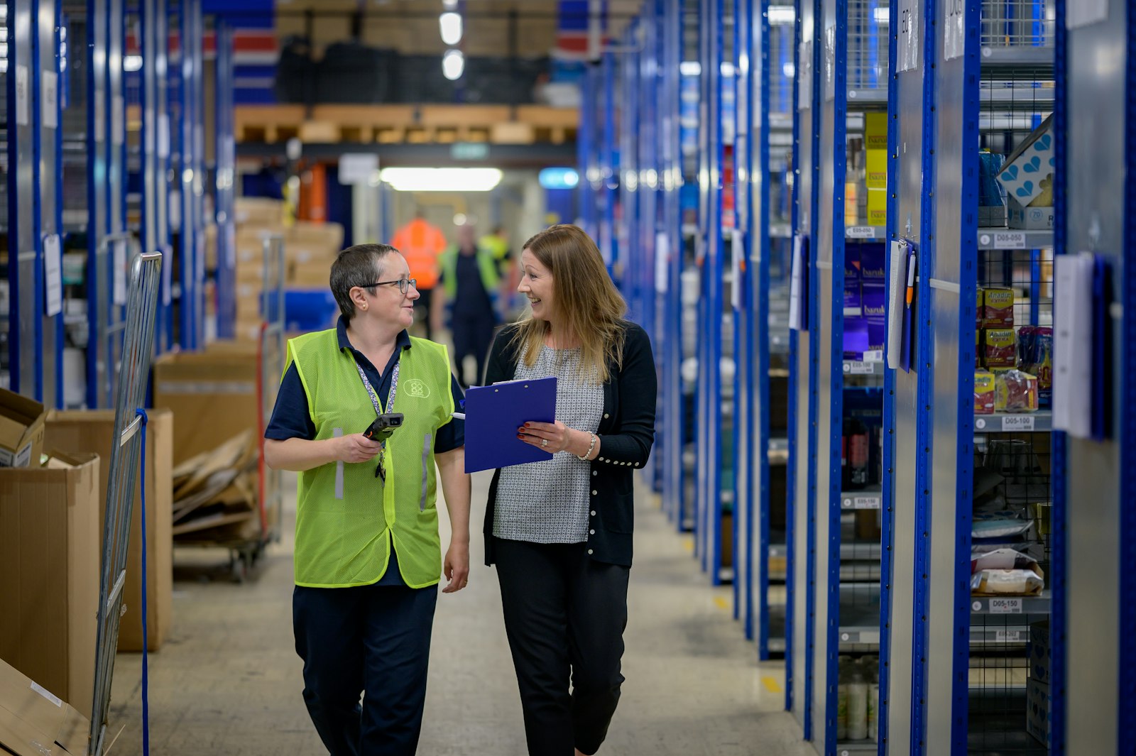 Two workers walking down a tall blue racking aisle in a warehouse, one in a yellow-green hi-vis vest holding a handheld scanner and the other carrying a clipboard