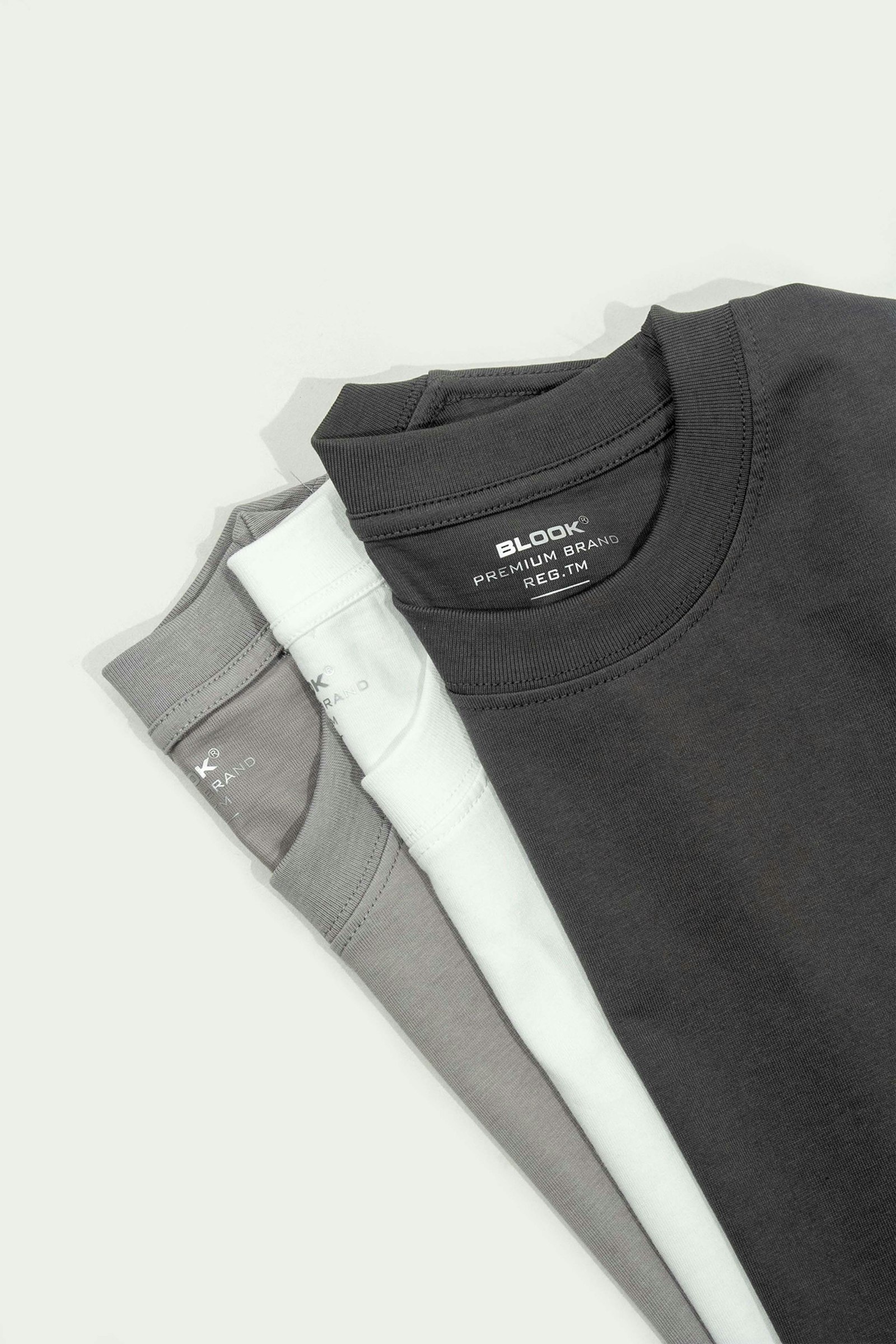 Three folded t-shirts in grey, white and charcoal stacked on a pale surface