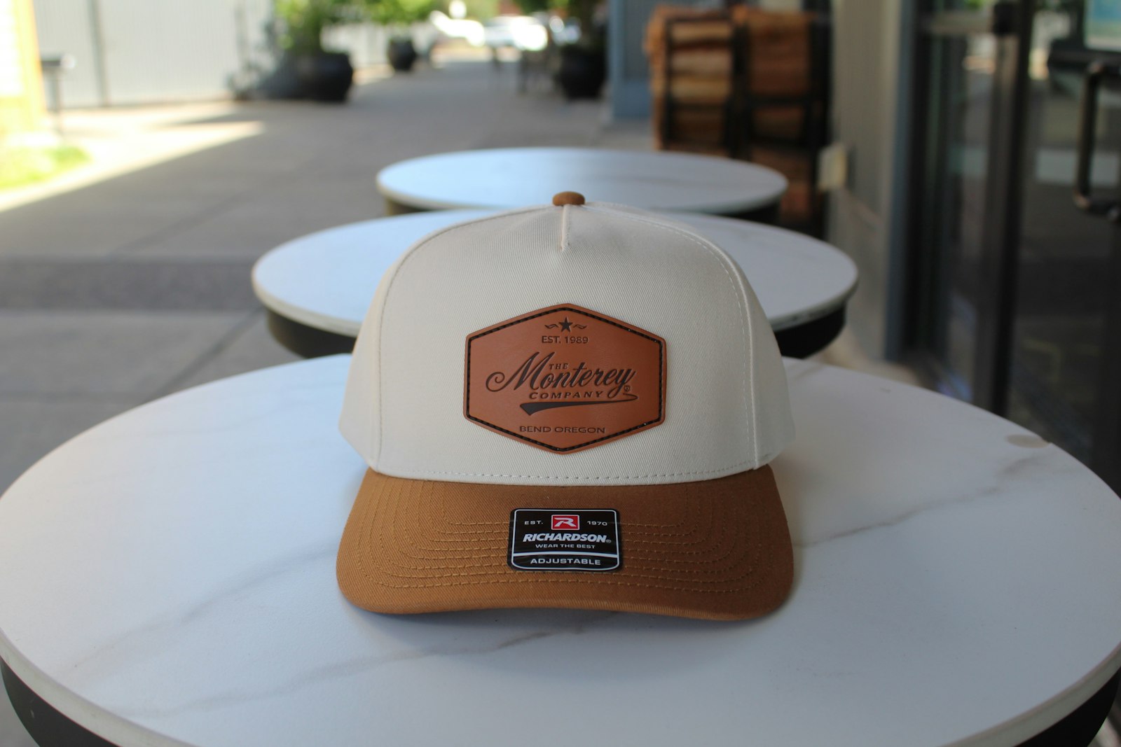 A cream and tan trucker cap with a stitched leather patch on the front panel, on a white table