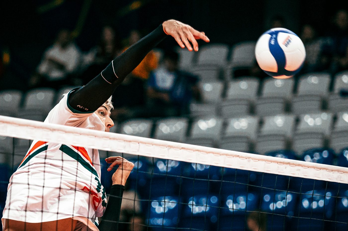 An indoor volleyball player in a white jersey with orange and green stripes spiking the ball over the net in front of near-empty arena seats