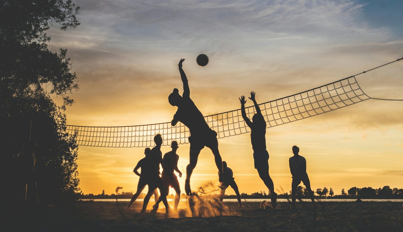 Silhouettes of people playing beach volleyball at sunset, one leaping to spike over a rope net against an orange sky
