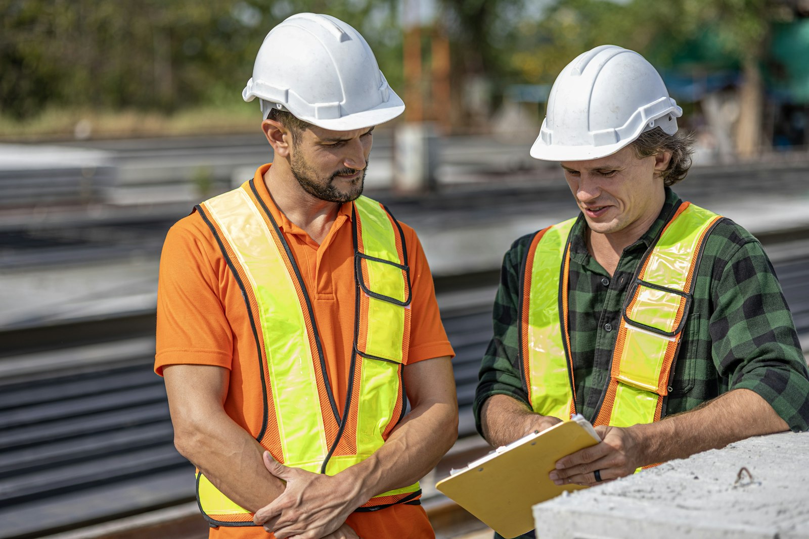 Two workers in white hard hats and hi-vis vests reviewing plans on a clipboard beside stacked concrete beams