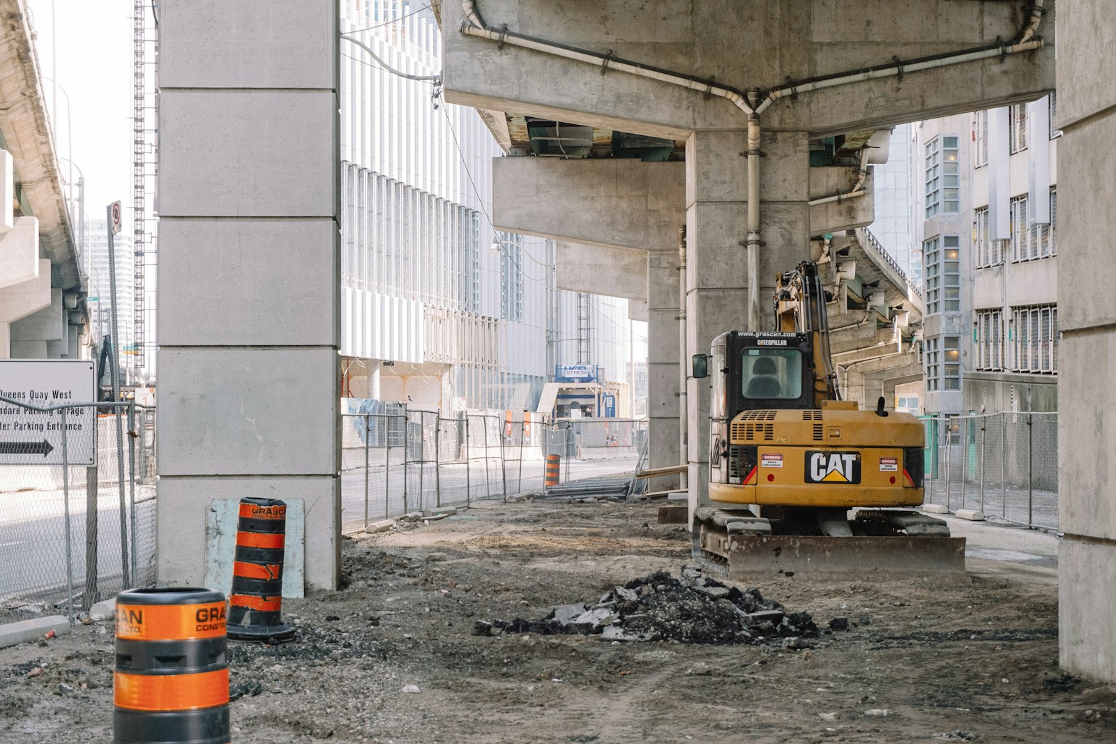 A construction site beneath an elevated concrete highway in downtown Toronto, with an excavator, orange-and-black traffic barrels and a chain-link fence