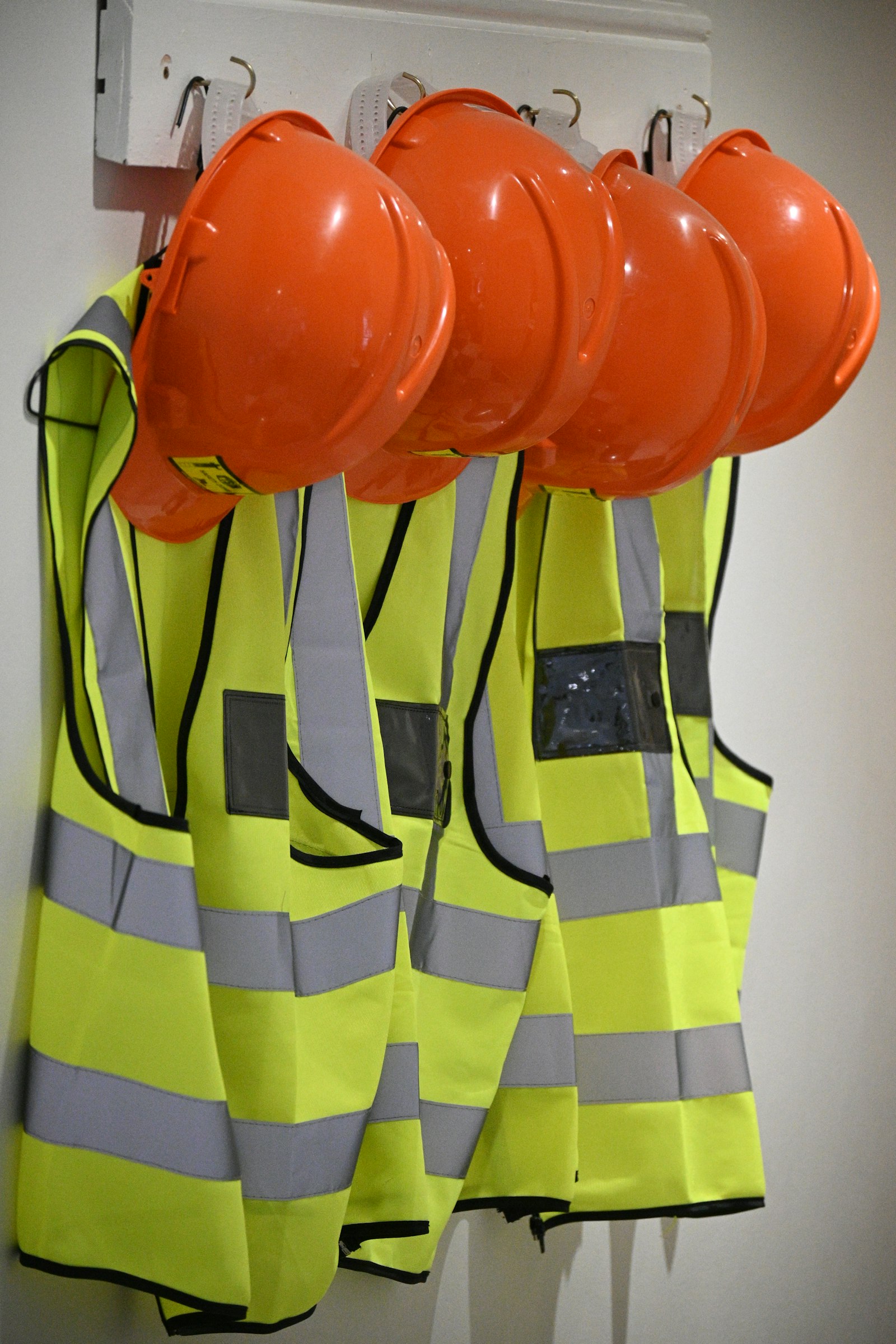 Four orange hard hats hung above four yellow hi-vis vests on a white wall