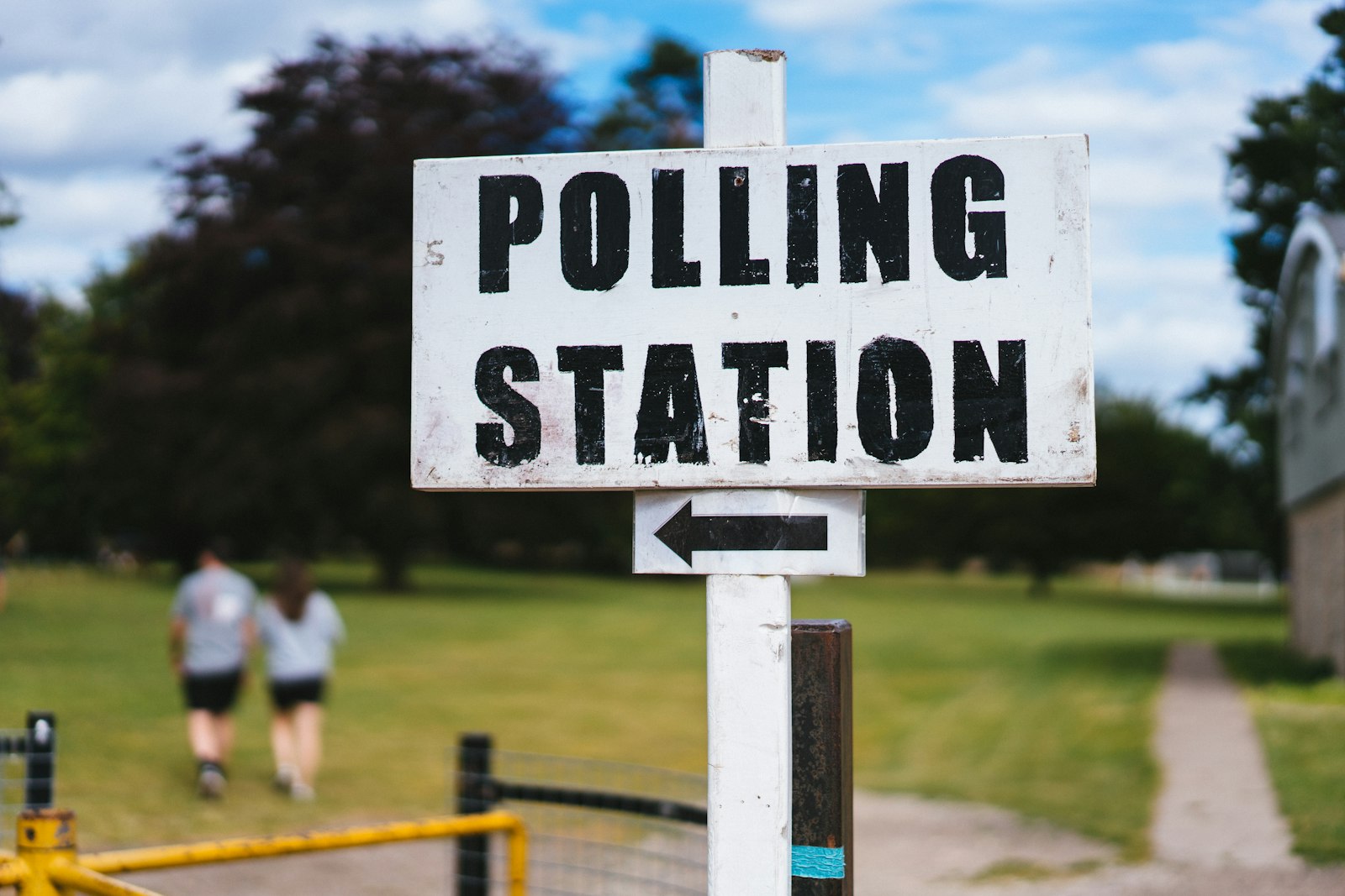 A white Polling Station sign with an arrow on a post in a park, with two people walking away in the background