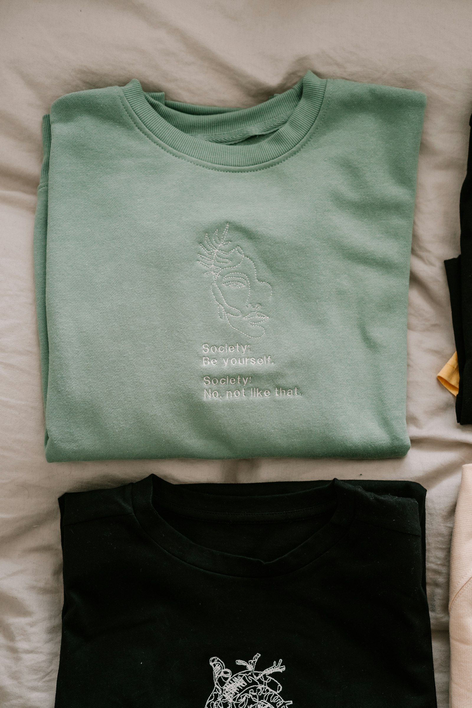 A folded pale green crew-neck sweatshirt with small embroidered detail, beside a black one
