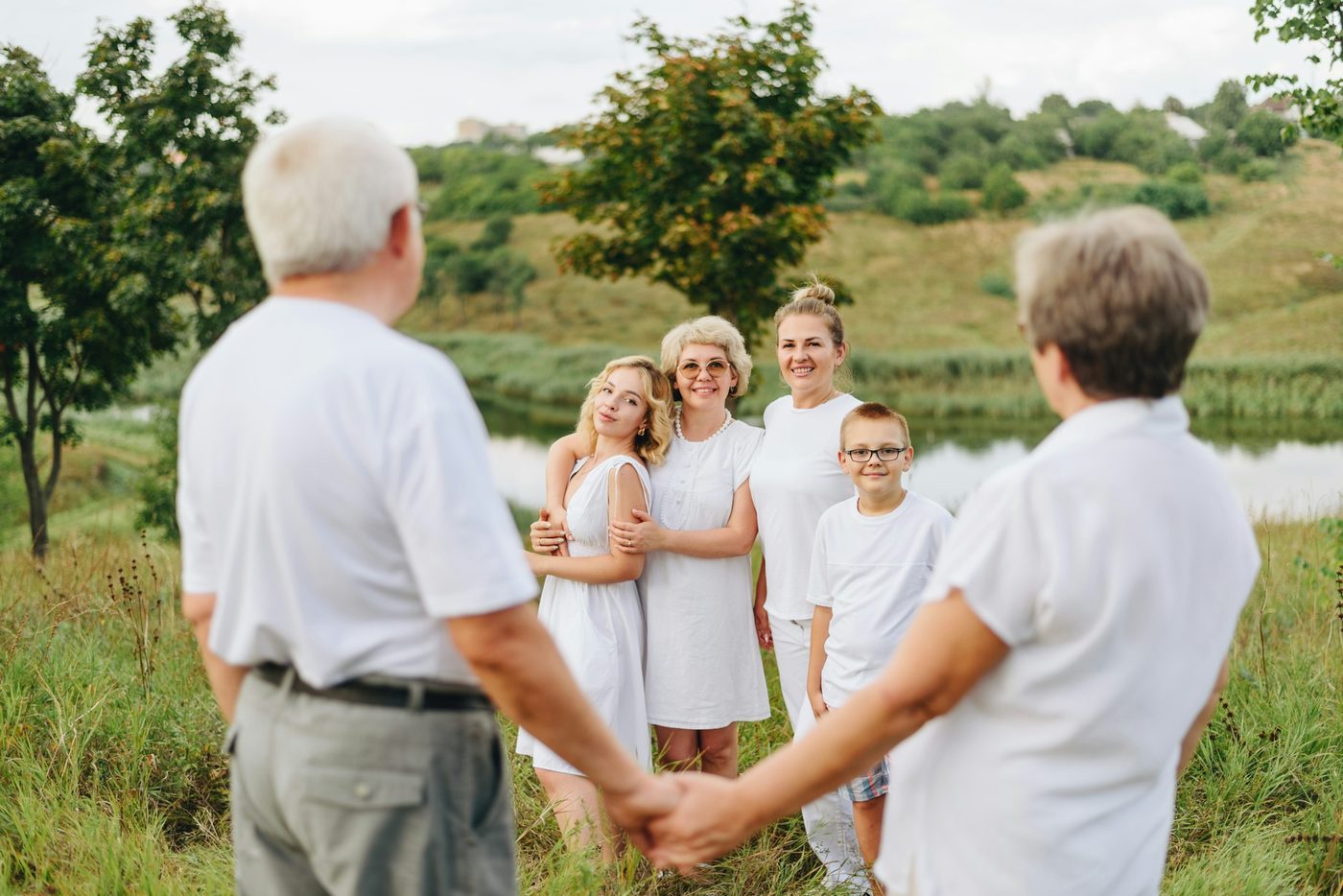 Grandparents in white seen from behind, holding hands and facing their daughters and grandchildren, all dressed in matching white, beside a pond