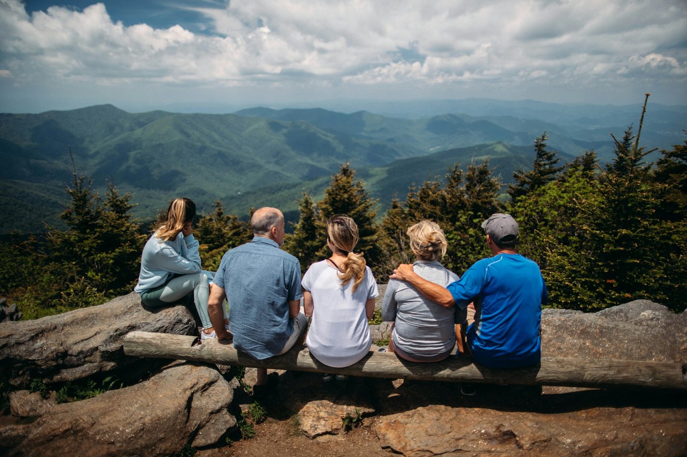 Five family members seen from behind sitting on a log rail at a mountain overlook, looking out over layered green ridges