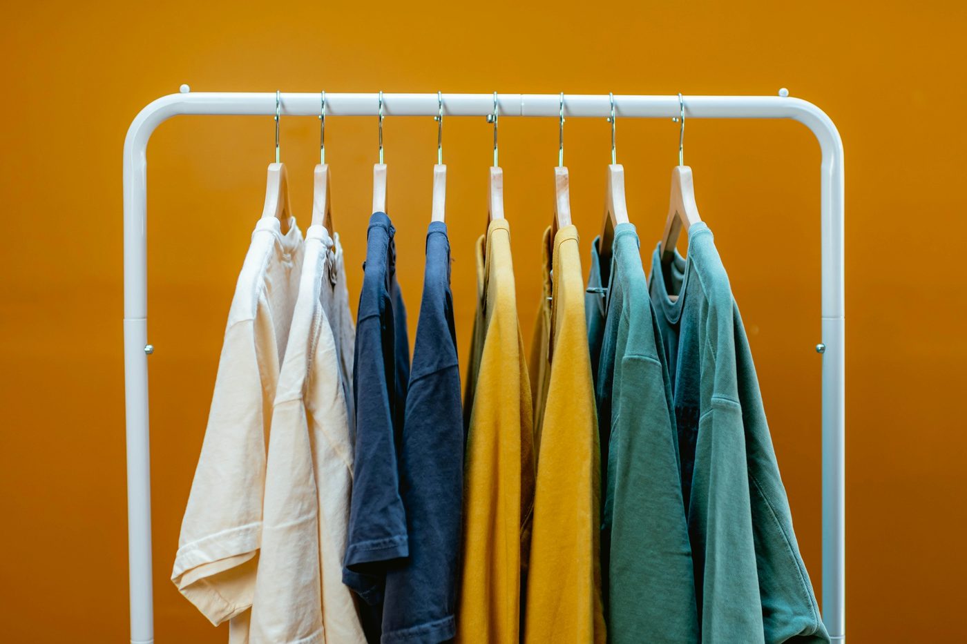 Eight plain t-shirts on wooden hangers in pairs of cream, navy, mustard and teal on a white rail against an orange wall