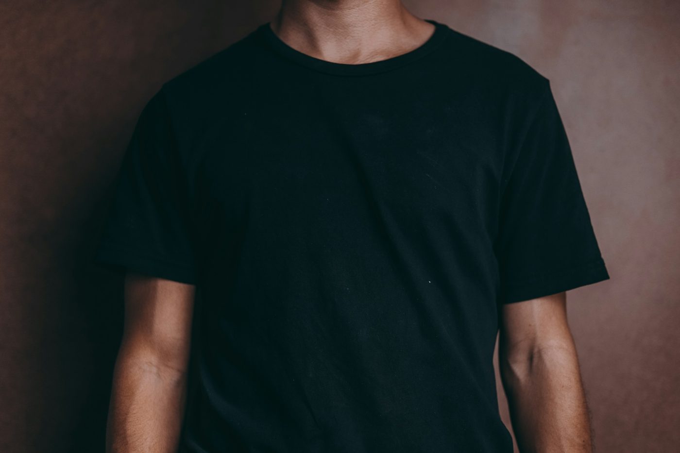 Torso of a man in a plain black crew-neck t-shirt, arms at his sides, against a warm brown wall