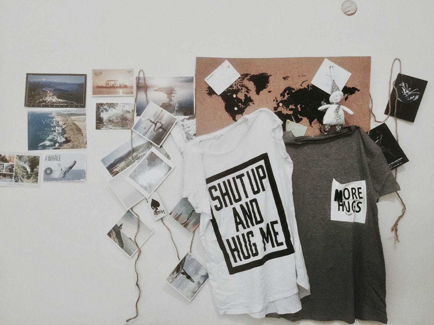 A white t-shirt with a large boxed “Shut up and hug me” front print beside a grey t-shirt with a small square “More hugs” chest print, pinned to a wall with postcards