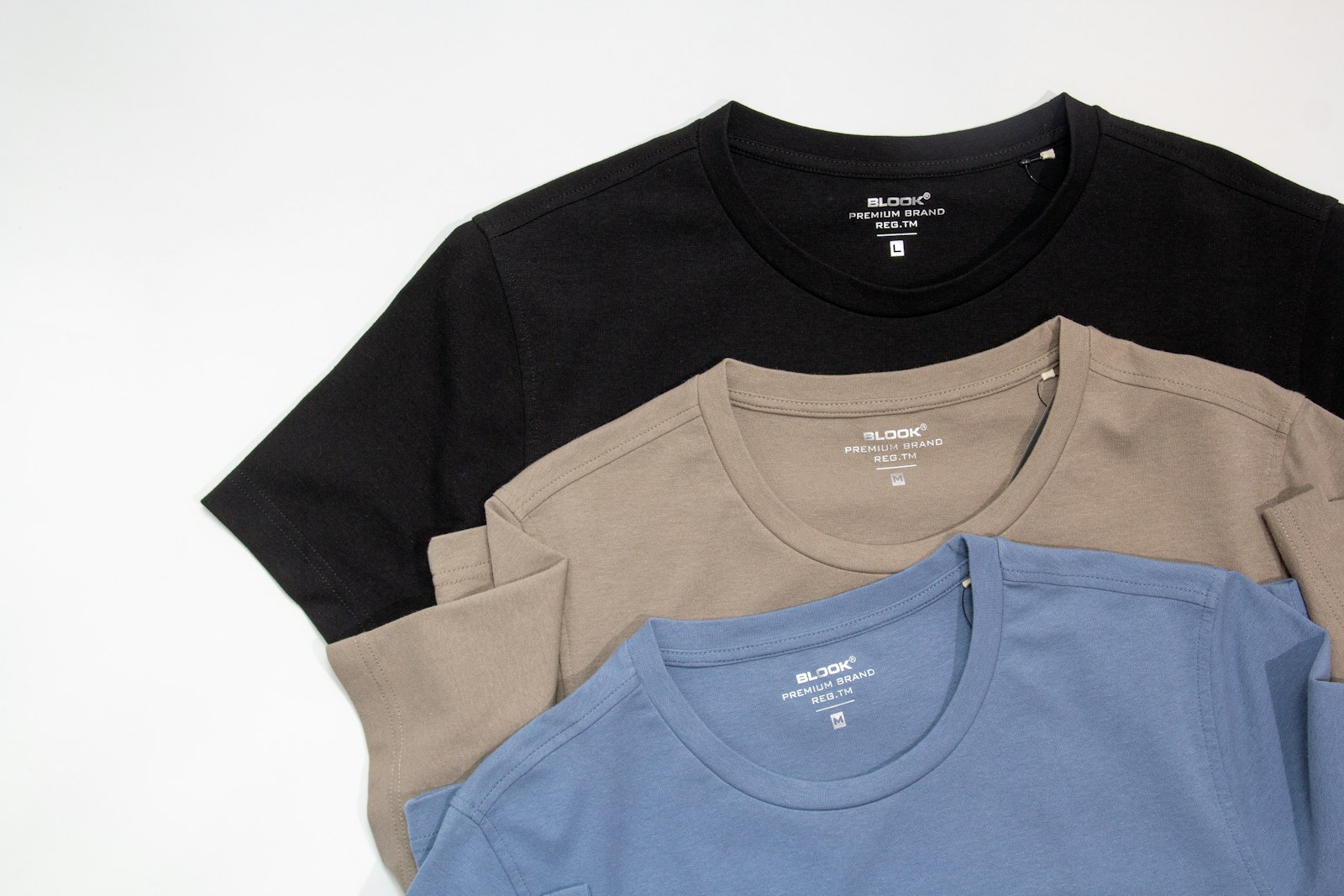 Three folded t-shirts in black, tan and blue stacked on a white surface