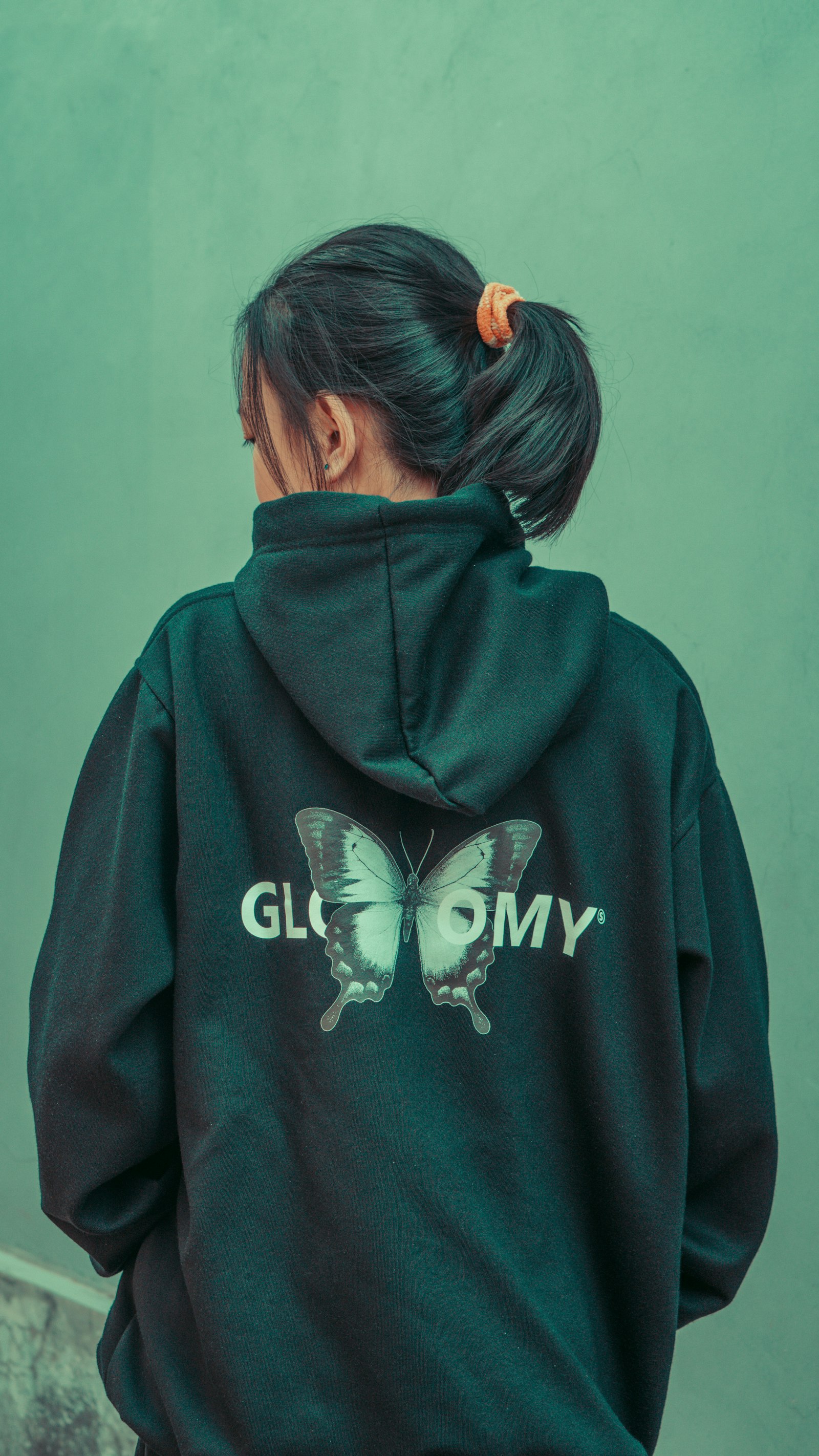 The back of a green hoodie with a printed graphic and lettering, worn against a teal wall