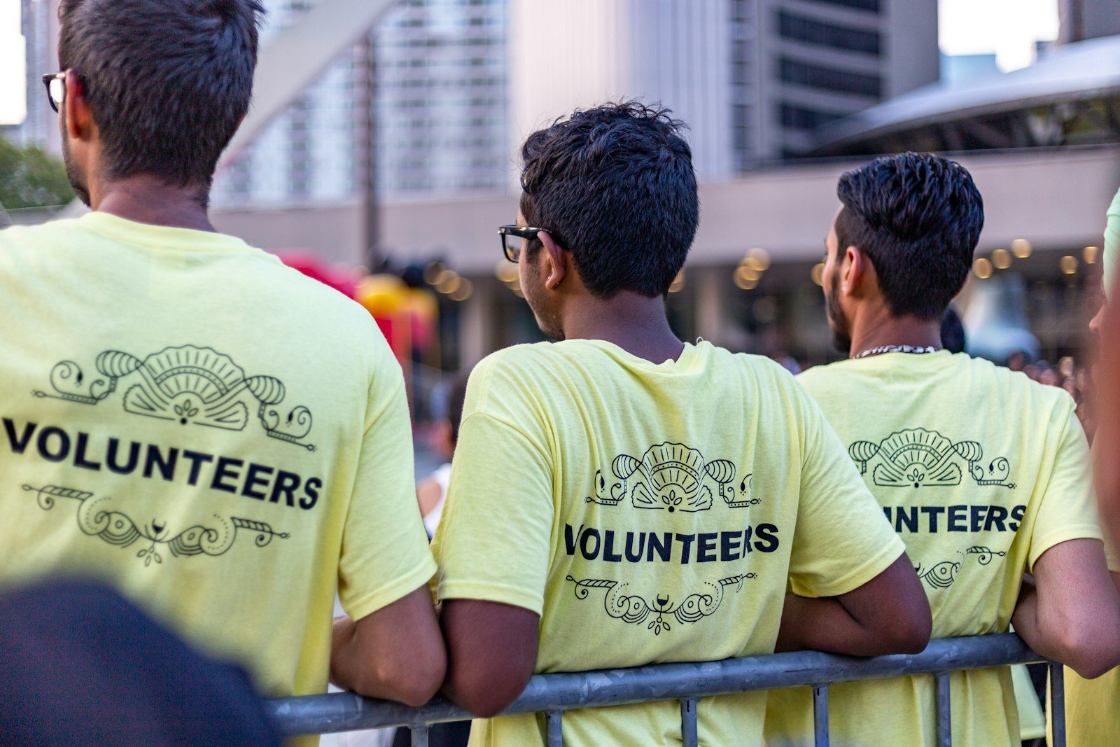 Three volunteers in matching yellow VOLUNTEERS t-shirts leaning on a barrier, seen from behind