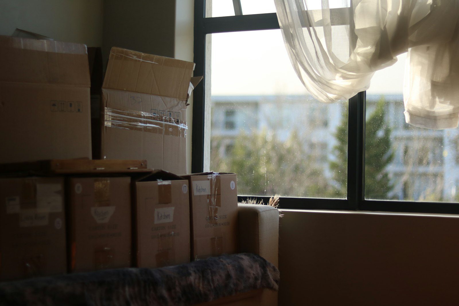 Cardboard boxes stacked beside a window in a dim room, a sheer curtain lifting in the light