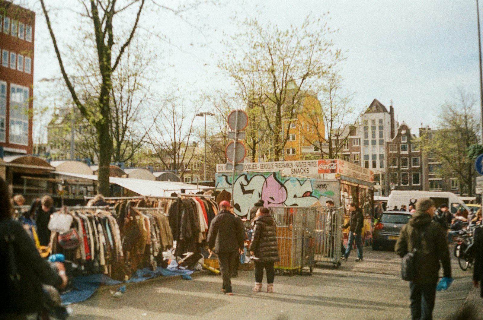 An open-air market street lined with clothing racks under canvas covers, shoppers browsing between the stalls