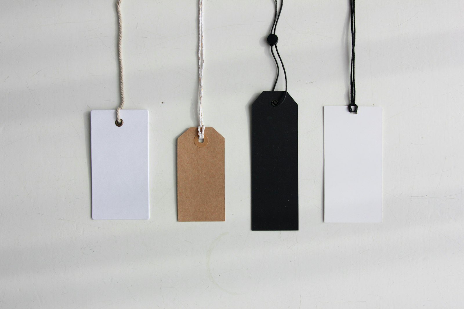 Four blank hang tags in white, kraft and black hanging from strings against a white wall