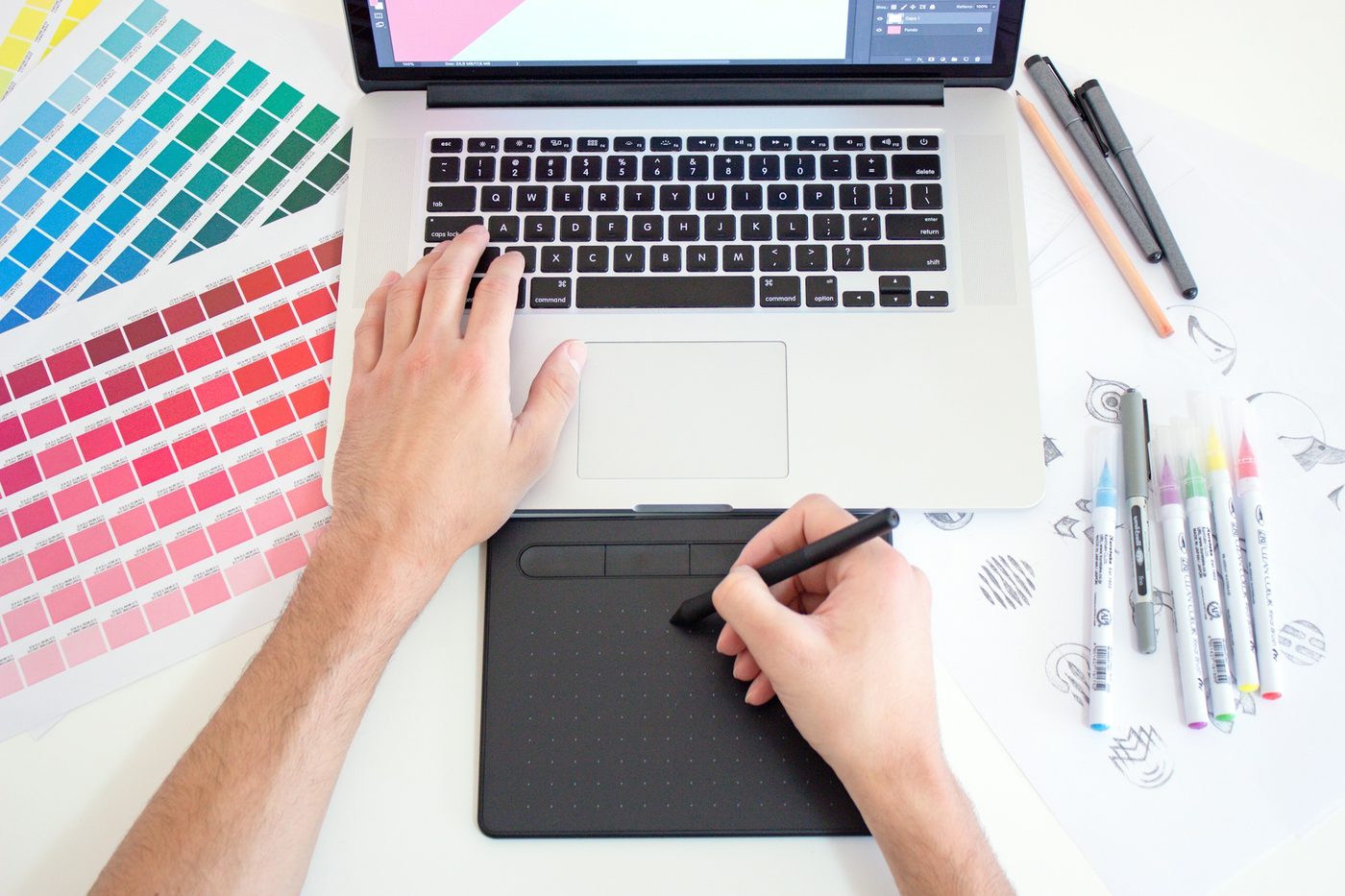 Overhead view of a designer’s hands on a laptop keyboard and a pen tablet, surrounded by colour swatch sheets and markers