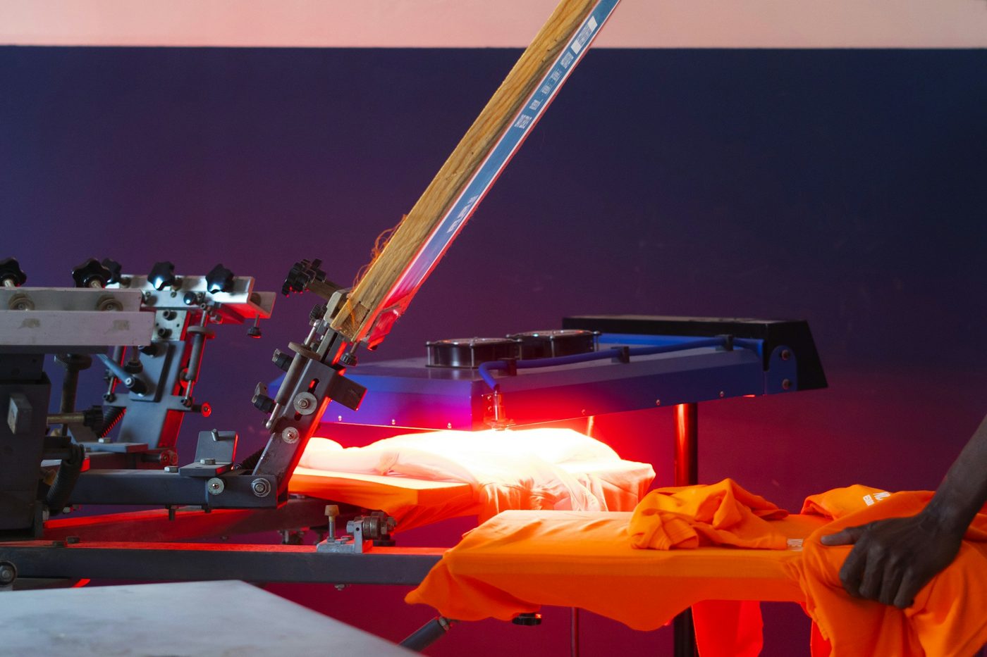 A manual carousel screen-printing press with a screen raised and a flash-cure unit glowing red over a garment, orange t-shirts on the next platen