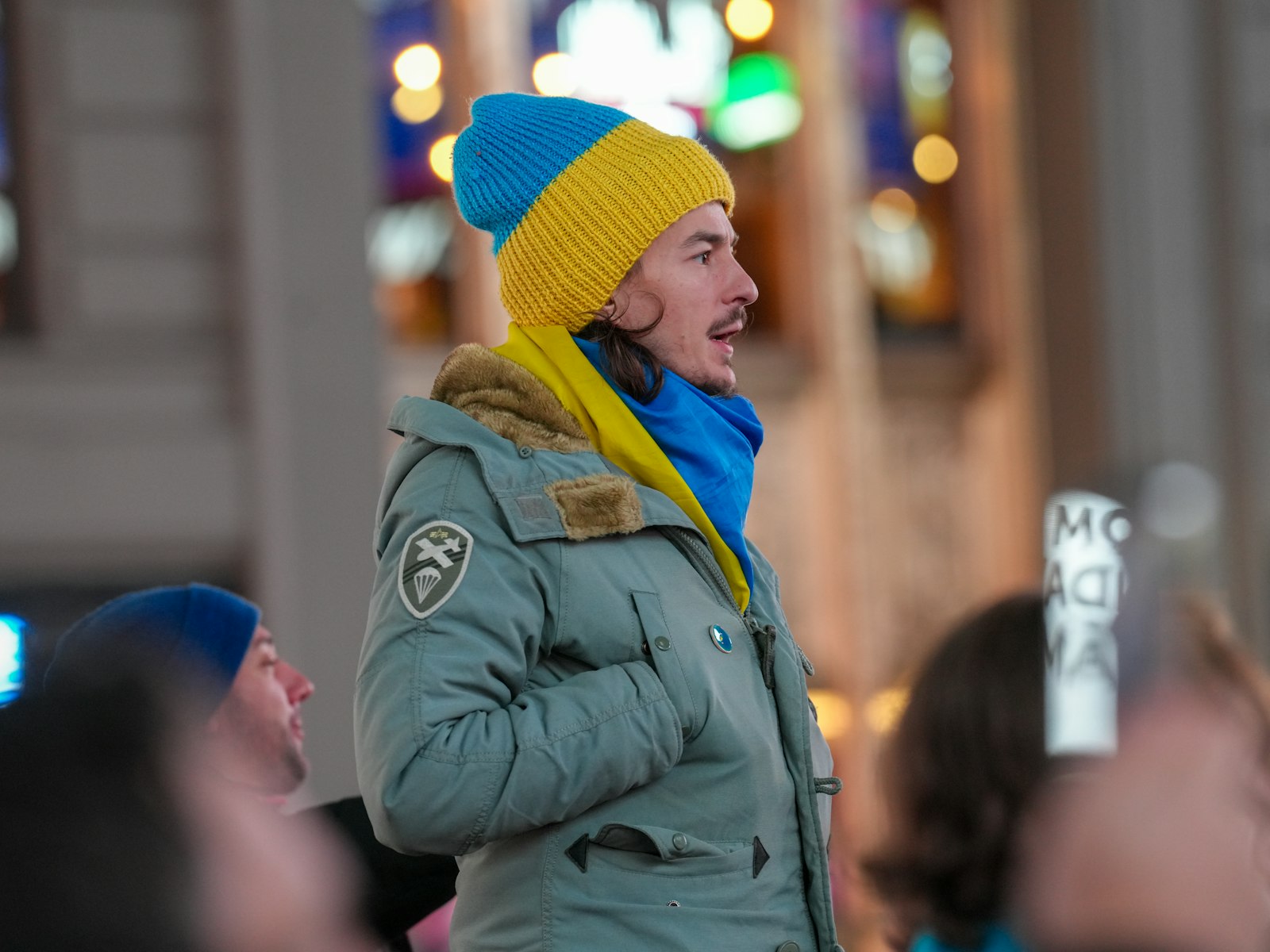 A person in a green insulated parka and a blue and yellow knit hat on a city street