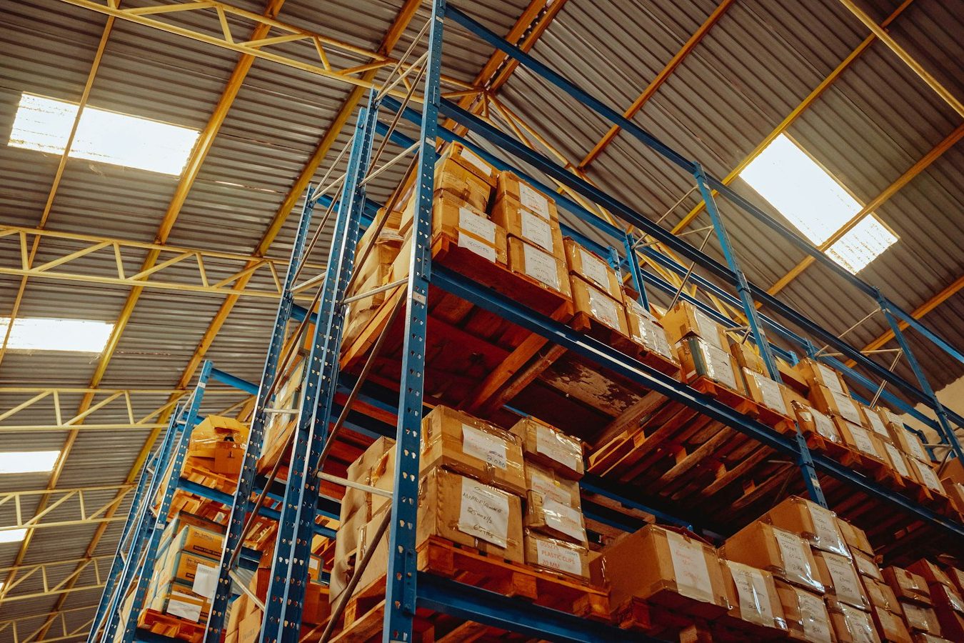 Pallets of cartons on tall blue warehouse racking, seen from below under skylights